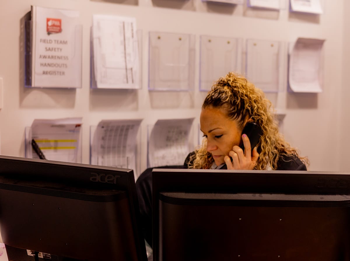 Office staff member at PWA Electrical Services speaking on the phone behind dual computer monitors, with safety documents and field team registers displayed on the wall