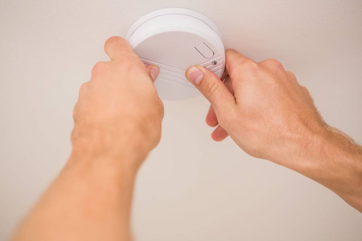 Close-up of a person’s hands installing or testing a smoke detector on a white ceiling.