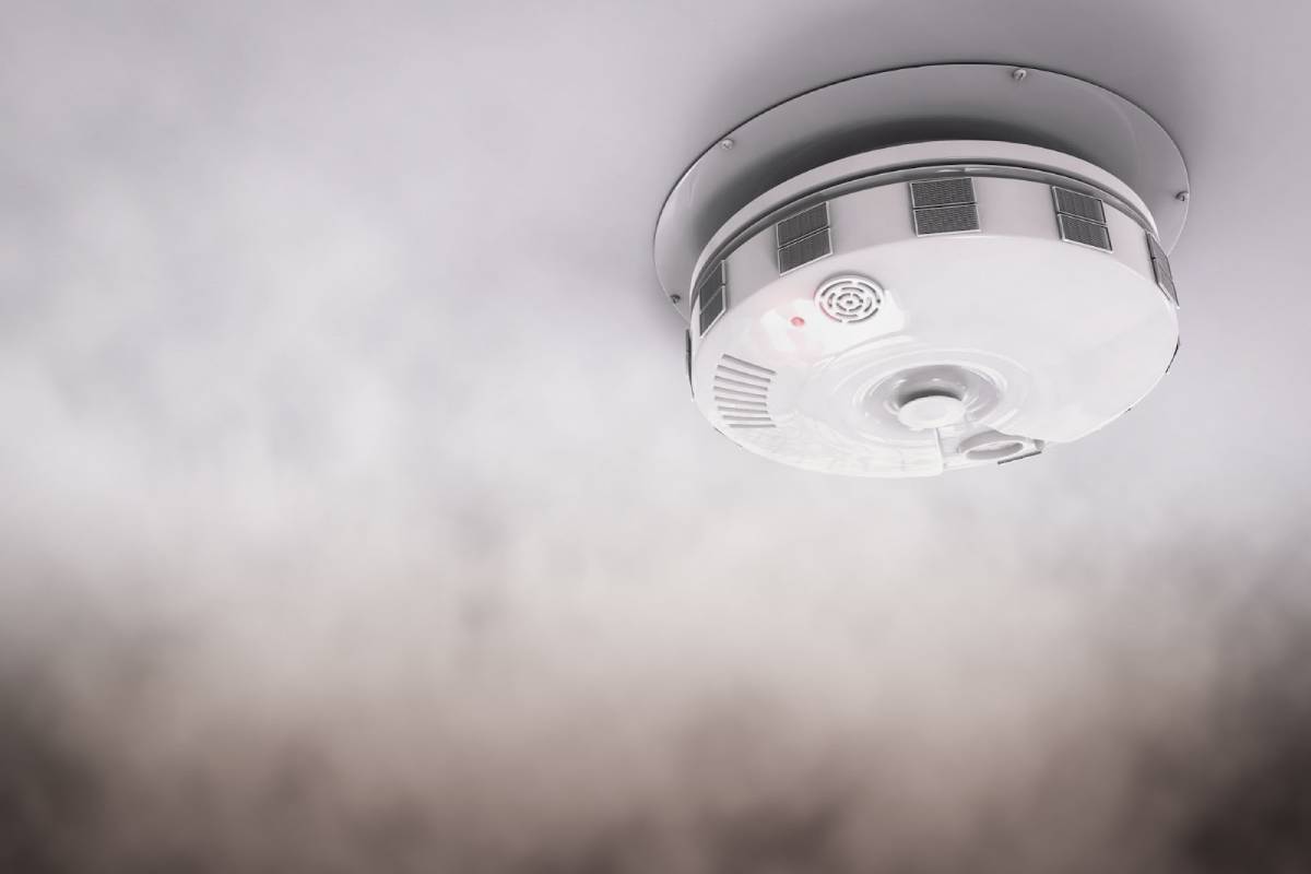 Close-up of a smoke alarm on the ceiling surrounded by smoke, with a red indicator light activated.