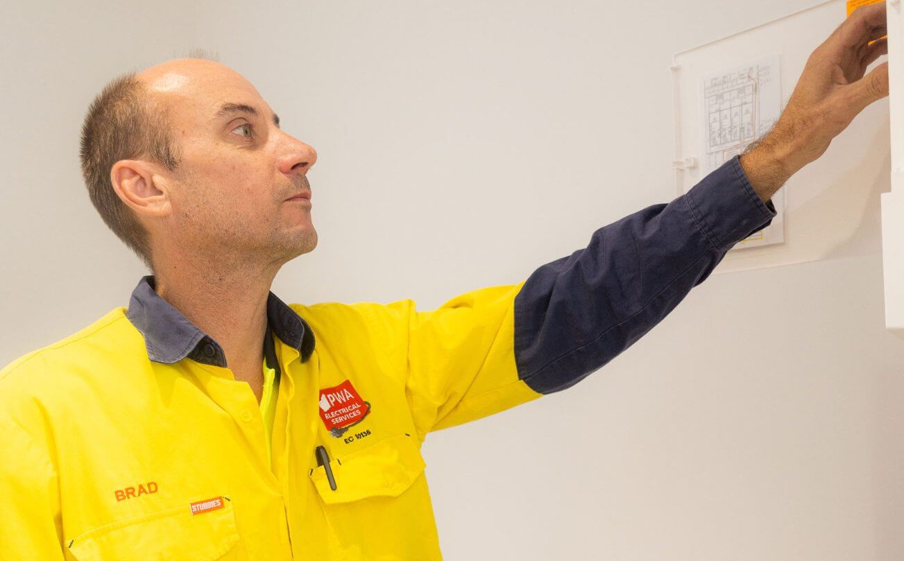 PWA Electrical Services technician inspecting a mounted electrical plan in a white-walled room, wearing a yellow and navy work shirt