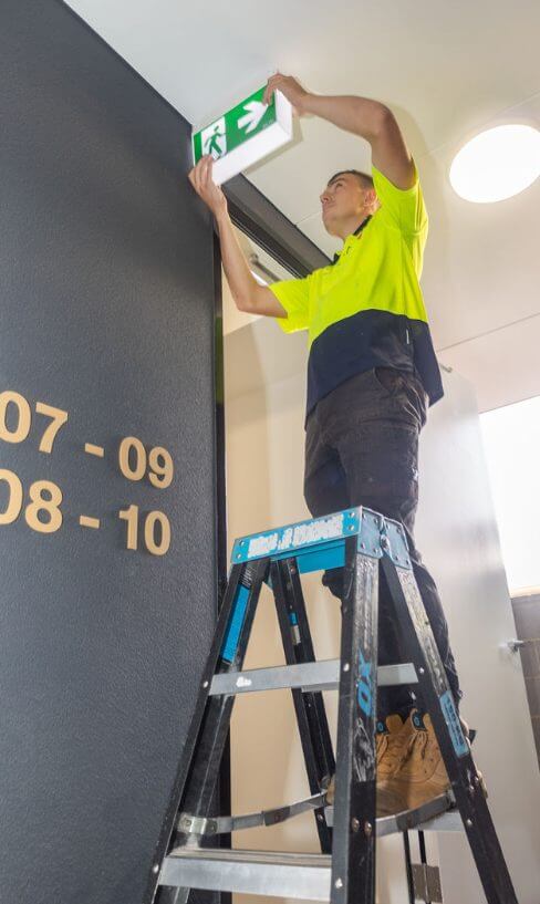 Electrician on a ladder installing an illuminated emergency exit sign above a doorway in a modern commercial building