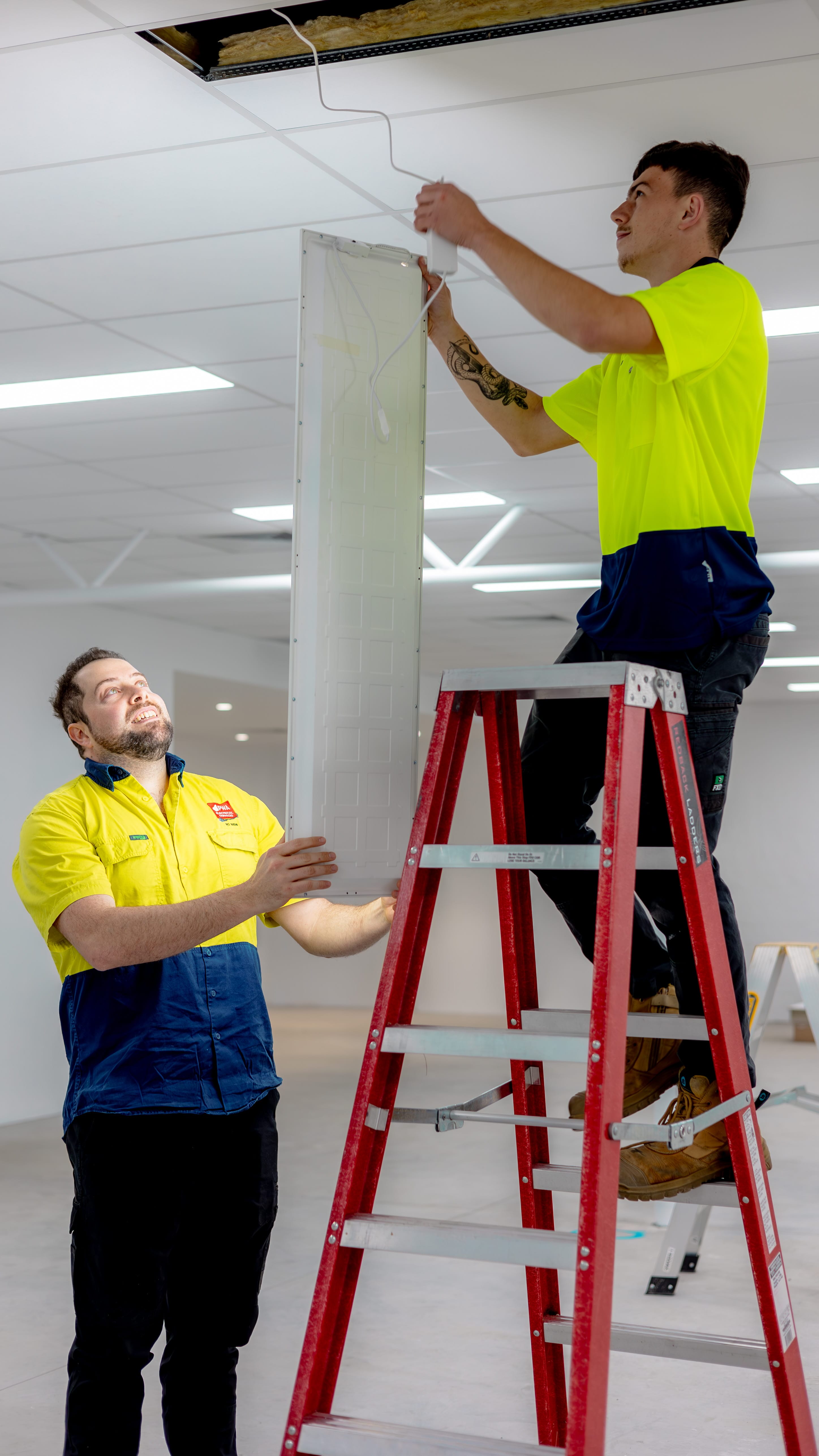 Electricians installing LED panel lighting in a commercial ceiling using a ladder.