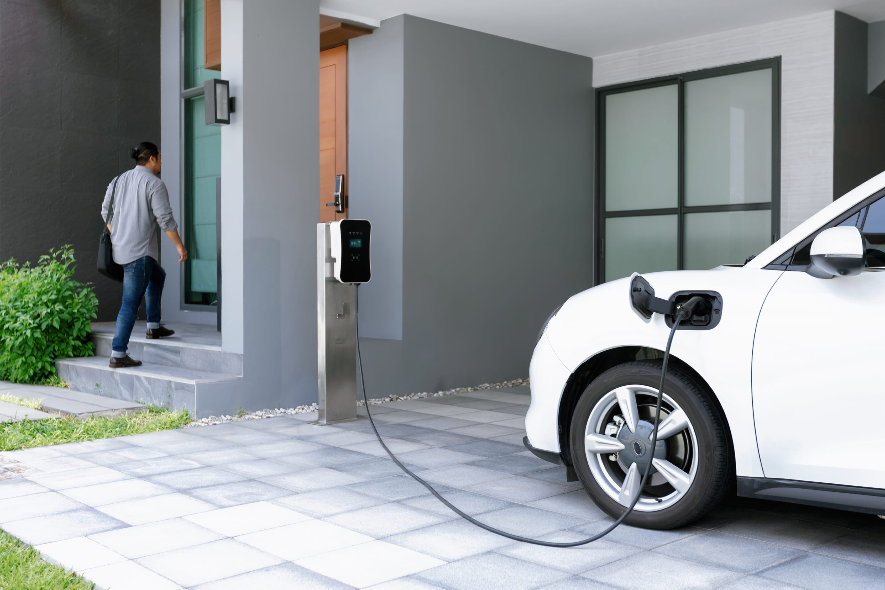 Electric vehicle connected to a home charging station with a resident walking into the house.