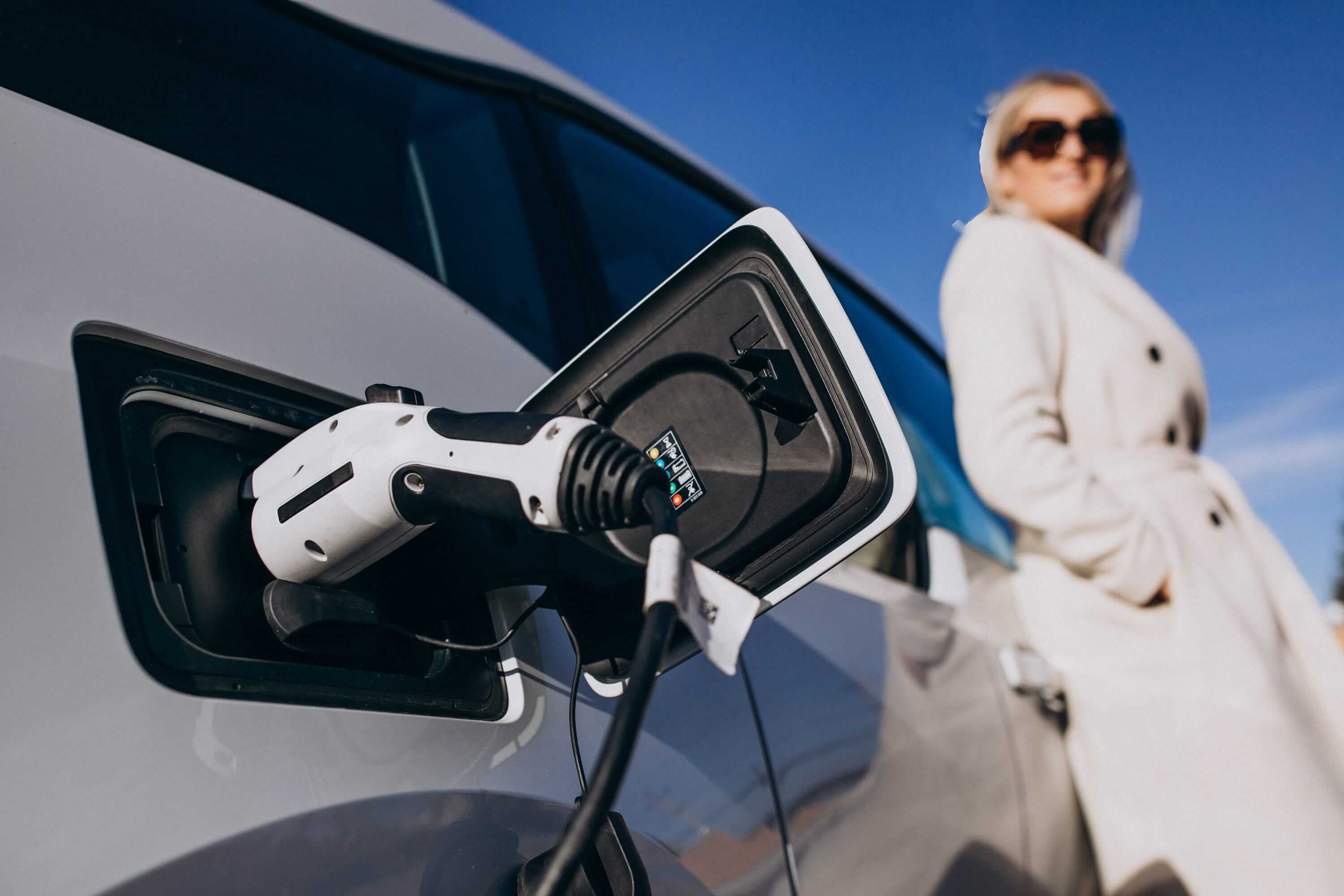 Woman standing near an electric vehicle charging at a home charging station outdoors.